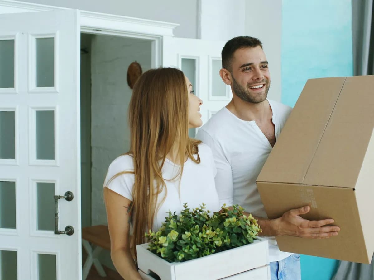 Couple carrying a moving box and a plant through the front door of their new home