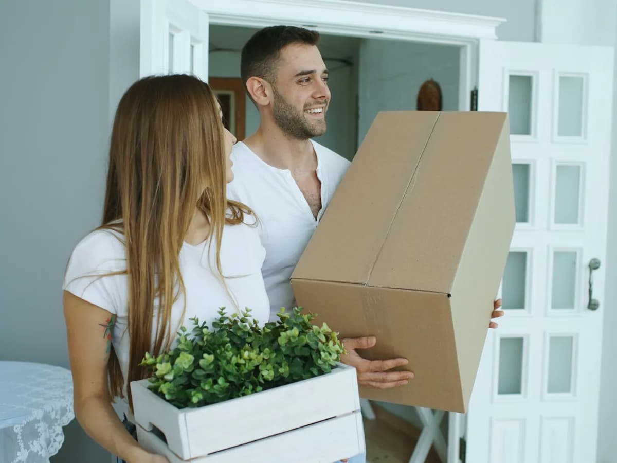 A couple carrying a moving box and a crate of plants into their home