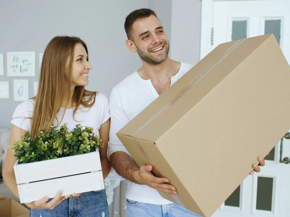 A couple carrying a large moving box and a plant into their new home